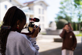 Une jeune personne qu'on voit de dos à gauche de la photo tient une caméra dans sa main et filme une autre jeune personne qu'on voit en arrière plan à droite. Elles se trouvent sur une place extérieure ensoleillée.